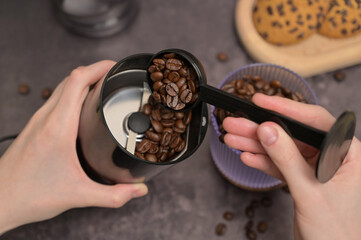 Close up. A woman's hands pours coffee beans into a coffee grinder from a transparent glass with a black spoon. Oatmeal cookies with chocolate chips on a wooden board. Dark background
