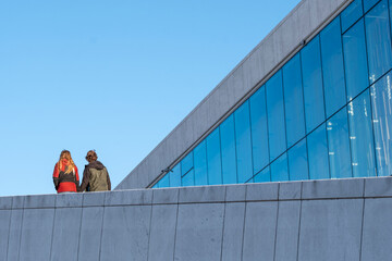 Two people walking along minimalist concrete terrace beside glass facade with clear blue sky showing human connection light and movement in modern Oslo architecture