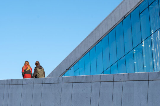 Two people walking along minimalist concrete terrace beside glass facade with clear blue sky showing human connection light and movement in modern Oslo architecture