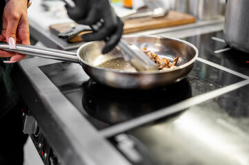 Chef cooking meat or mushrooms in stainless steel pan on electric stove in modern kitchen. Black glove, manicured nails, and utensils suggest professional food preparation.