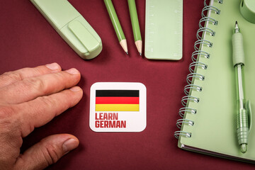 Hand next to Learn German card with Germany flag surrounded by stationery and notebook, concept of studying foreign language and education