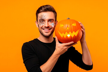 Man with Halloween makeup holding carved Jack-o'-lantern against orange background. Model in studio for Halloween celebration