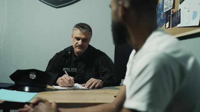 Police officer interrogating male witness in investigation room, taking notes