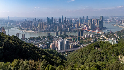A scenic view of the modern Chongqing skyline seen from a lush, green mountain overlook. The...
