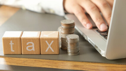Tax planning with stacked coins and wooden blocks spelling tax beside person using laptop, symbolizing financial management and online tax filing