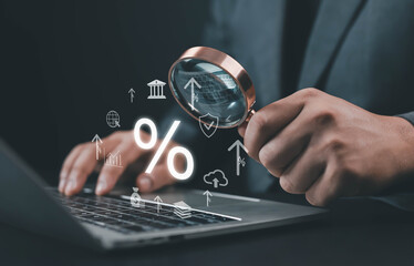 Businessman analyzing financial data and interest rates through a magnifying glass while using a laptop. Concept of banking, investment research, economy growth, and digital finance strategy.