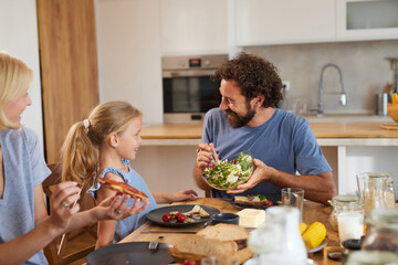A family shares a delightful dinner at home, with a father playfully serving a salad to his smiling daughter while the mother enjoys a plate of food, creating a warm atmosphere.