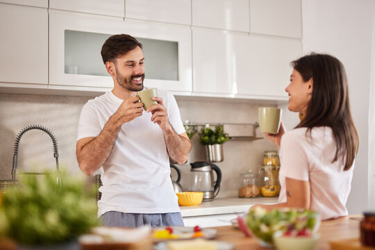 A couple shares joyful moments over cups of coffee in their stylish kitchen, surrounded by fresh ingredients and cheerful decor. They engage in light conversation and smiles.