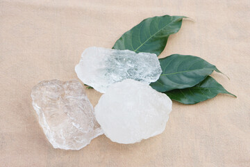 Natural alum crystal stones placed on green leaves over beige fabric background. The clear mineral blocks shine with purity and simplicity, representing eco wellness, spa, natural hygiene concept.    
