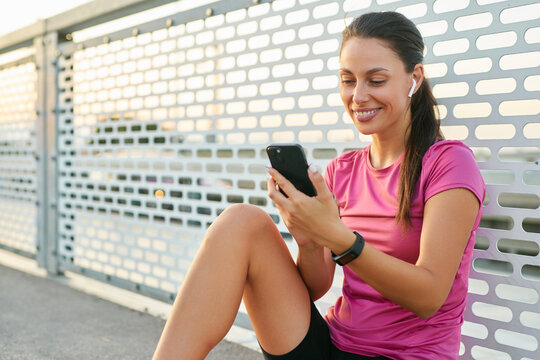 A woman enjoys her phone while resting on a bridge after an energizing workout. She wears athletic clothing and earbuds, with warm evening light enhancing the moment.