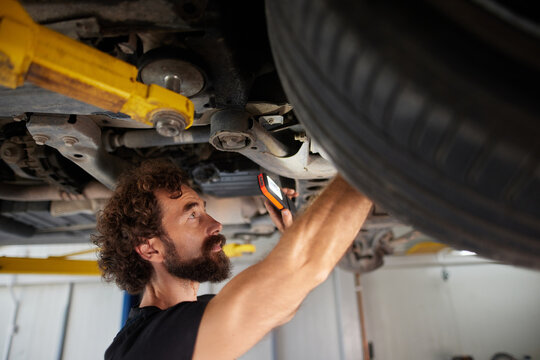 A mechanic is examining the undercarriage of a vehicle inside an auto repair shop, using a flashlight to check for any issues. The workshop is well-lit and organized.