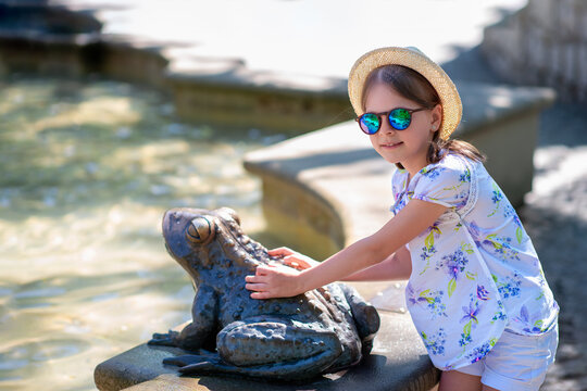 Girl in straw hat and floral shirt touching whimsical bronze frog statue by outdoor fountain in summer park, travel sightseeing and happy vacation moment, Swieradow-Zdroj, Poland