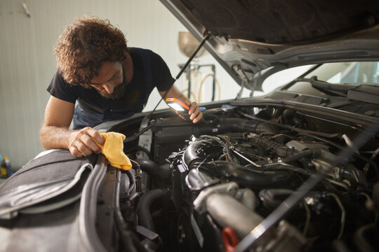 A skilled car mechanic inspects various parts of an engine using a flashlight while cleaning with a cloth, focused on his work in a bright repair shop.