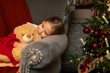 Christmas eve holiday. Charming little girl in red festive dress lying on chair, hugging teddy bear...
