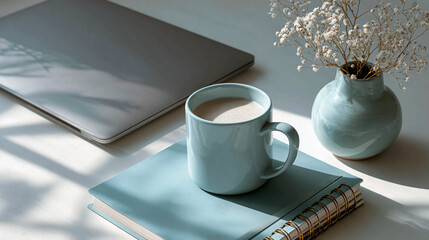 A workspace with a laptop on the left and a coffee mug in the center. There is a vase and a notebook on the right.