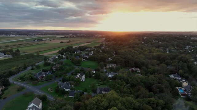 Luxury suburb neighborhood with large houses and swimming pool in garden. Rural farm fields and golden sunrise in distance. Aerial rising wide shot. American countryside city in Pennsylvania.