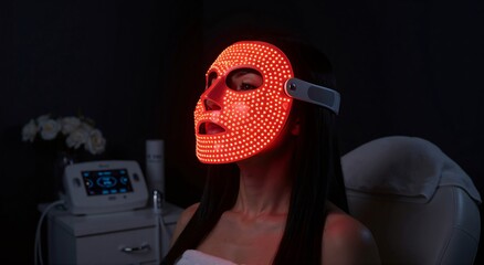 Woman getting LED light therapy on face wearing a red facial mask for skin rejuvenation. Cosmetology treatment at beauty clinic.