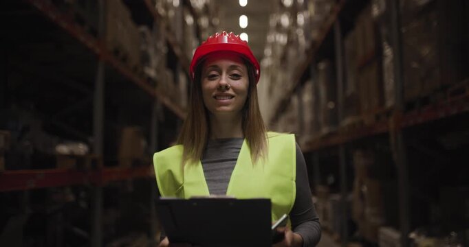 Confident female warehouse worker smiling with clipboard, wearing safety gear and looking at camera in industrial storage facility