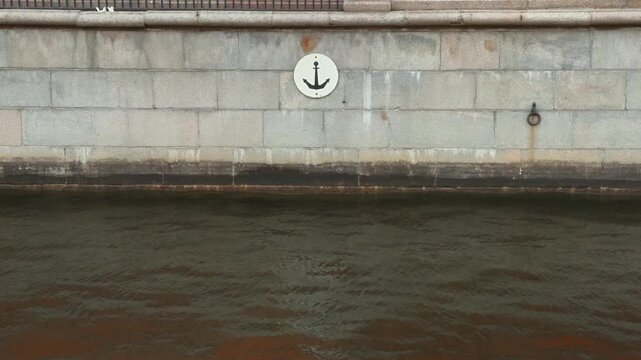 A close-up, straight-on shot of an old, pale-gray, massive granite block quay wall, featuring a white circular sign with a black anchor icon, positioned just above the dark, murky surface of a city