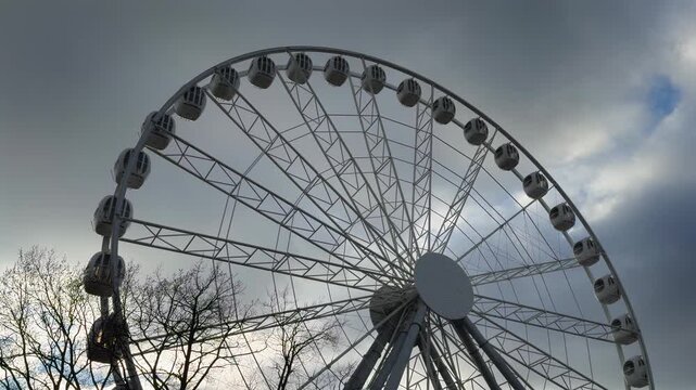 A panoramic, wide-angle, low-angle shot captures the top half of a massive, modern white Ferris wheel in a dramatic silhouette against a dark, stormy, overcast sky with patches of bright light and