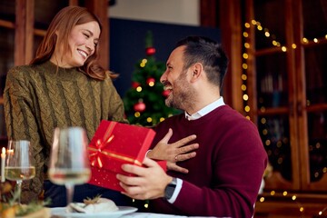 Happy Couple Share Gift Exchange During New Year Dinner Beside Christmas Tree In Cozy Home