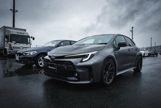 Matte gray Toyota Corolla GR Sport sits in a wet paddock with KRM truck and dark sedan, damp asphalt reflecting under overcast light at a Japanese racing facility.