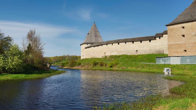 A panoramic, wide-angle shot of a calm river winding past an ancient, towering medieval fortress with light stone walls and a dark conical roof on the right. The left bank contrasts with a picturesque