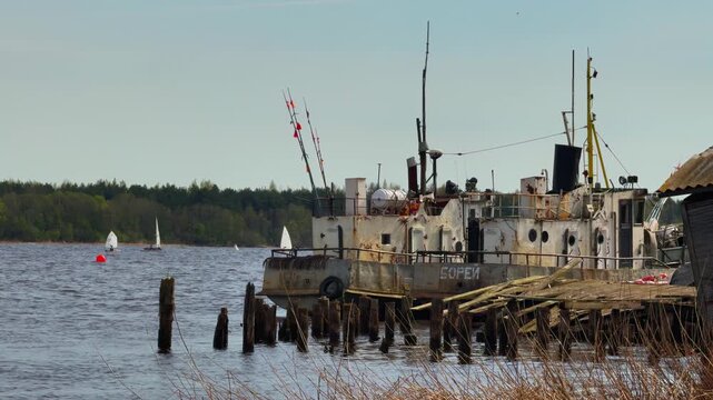 A mid-range, level shot of a large, heavily weathered and rusty white decommissioned cargo or service ship with the Cyrillic name Borey visible on its hull, docked next to a crumbling wooden pier on a