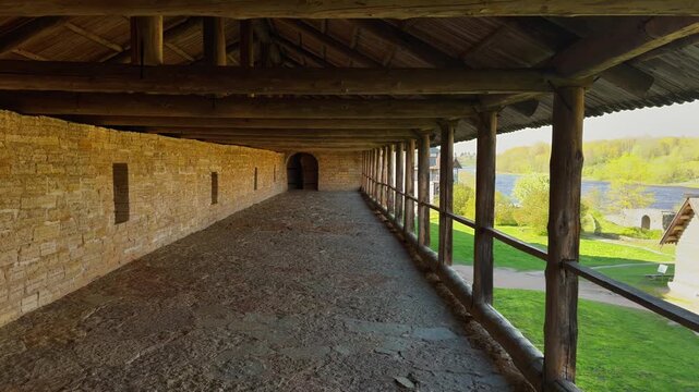 A medium, wide-angle shot of a long, empty covered walkway or gallery running along the inside of a medieval fortress wall made of light-colored, rough stone blocks. The roof and railing are supported