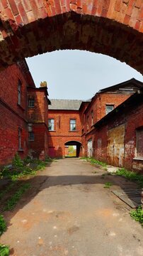 A medium, straight-on shot looking down a narrow, empty dirt and paved courtyard surrounded by tall, aged buildings of deep red brick. The sunlight highlights the weathered texture and peeling stucco
