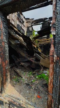 A close-up, straight-on shot of an empty wooden doorway, where the frame and log walls are intensely charred and blackened by fire. The doorway opens into the dramatic interior of the completely
