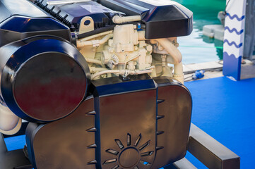 Detailed view of a large marine engine displayed on a black metal frame indoors, featuring black protective covers and white pipes, set against a blue floor background.