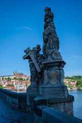 Fototapeta premium Sculpture of Socha svaté Anny on Charles Bridge over the Vltava River in Prague Old Town, Czech Republic, with Prague Castle visible in the distance under a clear blue sky.