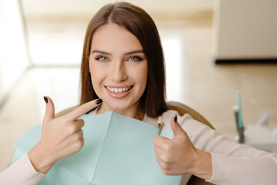 Smiling beautiful girl is sitting in dentistry chair with positive emotion and gesturing fingers up. Female patient visit dentist for regular teeth mouth cavity check up at medical clinic
