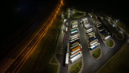 Aerial view of illuminated truck stop at night with parked vehicles along highway, providing...