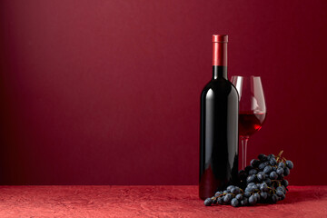 Bottle and glass of red wine with blue grapes on a red background.