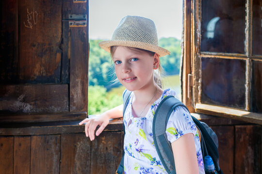 Smiling girl in straw hat and backpack standing by rustic wooden window with scenic lake and forest view, summer travel and sightseeing portrait outdoors