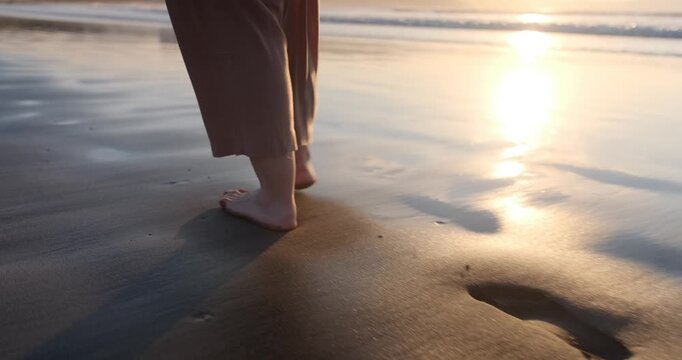 Slow motion close-up of a woman's bare feet walking on wet sand. The golden sun reflects on the shoreline as she leaves footprints.
