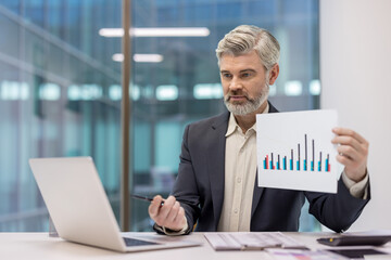 Mature businessman presenting colorful bar chart report during a virtual conference call, discussing financial data and business growth from a modern office with his laptop