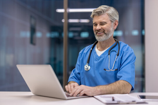 Experienced male doctor wearing scrubs and stethoscope, typing on a laptop with a cheerful expression, managing digital patient records in a healthcare environment