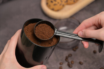 Close up. A woman's hand pours freshly ground coffee out of the coffee grinder with a black spoon. Oatmeal cookies with chocolate chips on a wooden board. Dark background