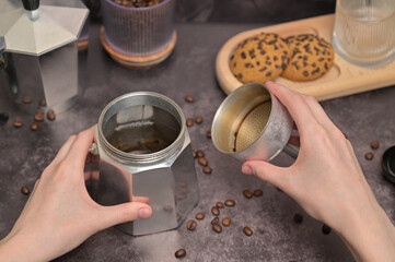 Close up. A woman's hands holding a geyser coffee maker with water. Coffee beans in a glass. Oatmeal cookies with chocolate chips on a wooden board. Dark background