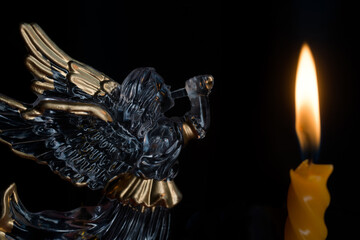 Glass angel with a trumpet beside a lit twist candle on black. Symbol of mourning for the deceased, prayer and memory in the dark, solemn vigil background. Photo