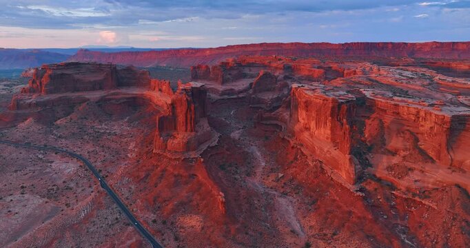 Red walls of the majestic canyons are lit with setting sun. Cars ride by the desert in the Arches National Park, Utah, USA. Aerial view.