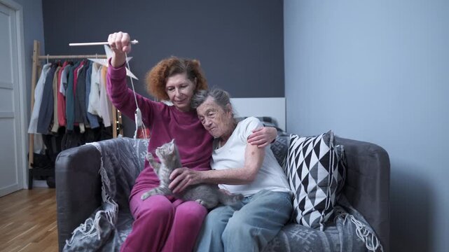 Volunteer assistant uses kitten to improve mood and physical condition of female senior patient at nursing home. Old grandmother and mature daughter playing with little cat toy sitting on sofa.