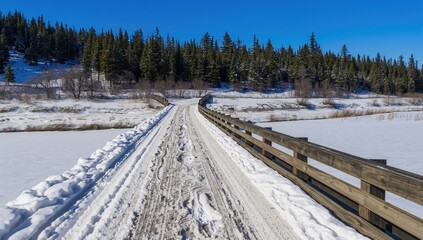 Obraz premium An aged wooden bridge spans a frozen stream, with tire impressions pressed into the snowy ground. Snow blankets the bare wooden railings, surrounded by evergreen trees on a hillside under a bright