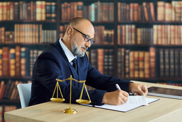 Lawyer signing contract document sitting at office desk of law firm with golden justice scales and...