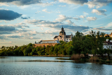 Obraz premium Beautiful view of an old monastery standing on a green hill above a calm river under a cloudy blue sky. Peaceful landscape with reflections on the water, trees, and historic architecture in warm eveni