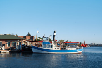 A blue and white passenger boat rests at a calm harbor beside brick quay buildings. Turkish flags fly as the vessel sits ready for tours in a sunny waterfront setting. © Loginova