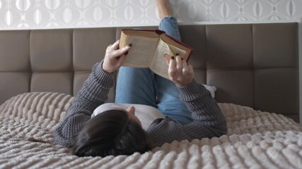 A young woman lying on bed and reading a book in a cozy bright bedroom. Peaceful domestic atmosphere, relaxation and self-care moment. - Powered by Adobe
